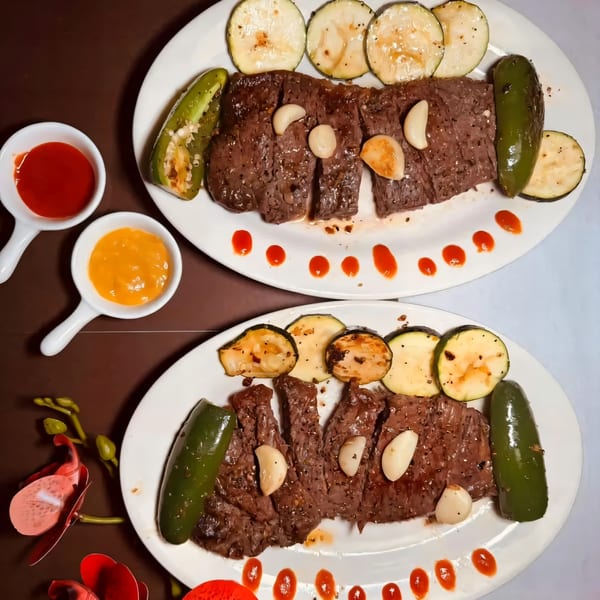 Two plates of sliced steak with roasted zucchini, jalapeños, garlic cloves, and dipping sauces.